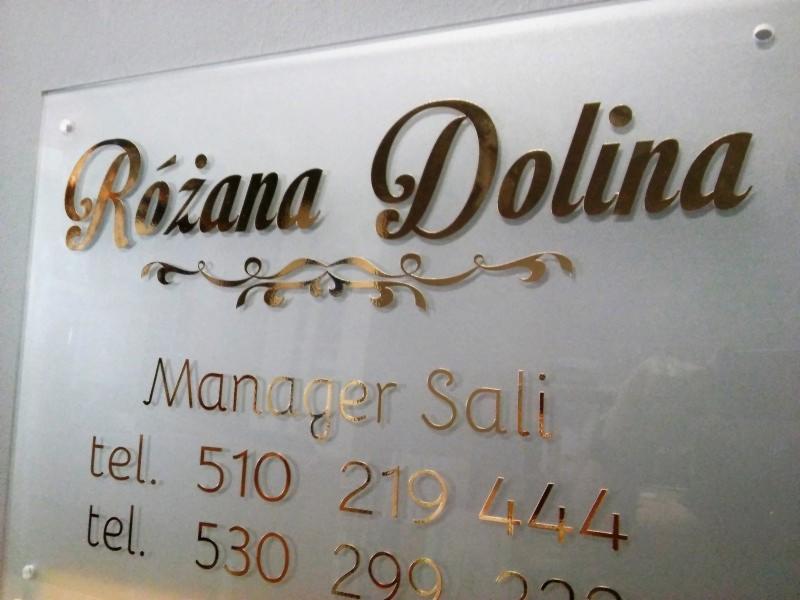 Szklana tabliczka z nazwą firmy 'Różana Dolina', imieniem managera i numerami telefonów, wykonana złotymi, ozdobnymi literami.