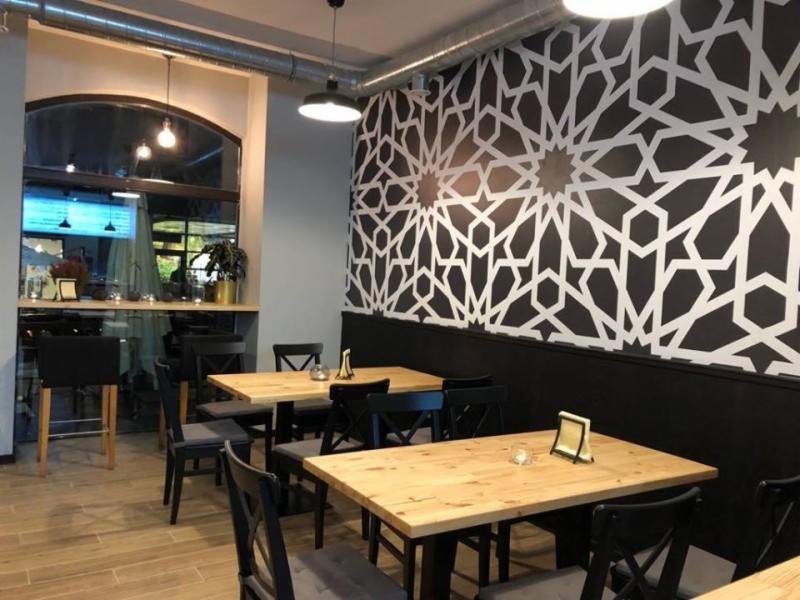 Wnętrze kawiarni z drewnianymi stołami i krzesłami, na ścianie dekoracyjny wzór geometryczny w kolorach czerni i bieli, oświetlenie punktowe.