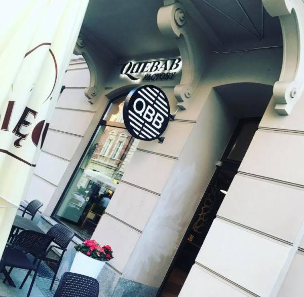 Elewacja budynku z szyldami 'QIEBAB FACTORY' i okrągłym logo 'QBB' z poziomymi paskami, widok z zewnątrz z stolikami i krzesłami kawiarnianymi oraz kwiatami w donicy.