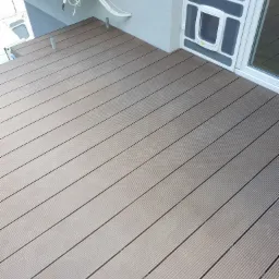 Nowoczesny taras z desek kompozytowych w odcieniach szarości, przylegający do jasnej elewacji z oknem i drzwiczkami dla zwierząt, widoczne przewody klimatyzacji na ścianie.