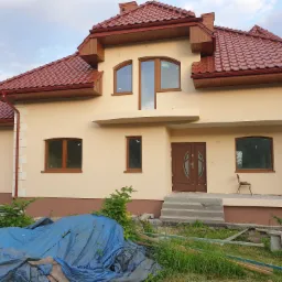 Dwukondygnacyjny dom z beżową elewacją, brązową dachówką i drewnianą stolarką okienną, z widocznymi śladami prac budowlanych na zewnątrz, w tym niebieska plandeka i resztki materiałów budowlanych.