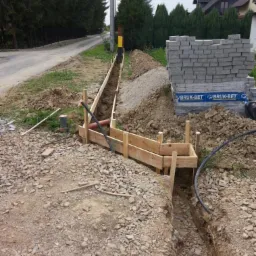 Przygotowanie wykopu pod fundament z drewnianym szalunkiem i ułożonymi rurami, obok paleta z kostką brukową Bruk-Bet.