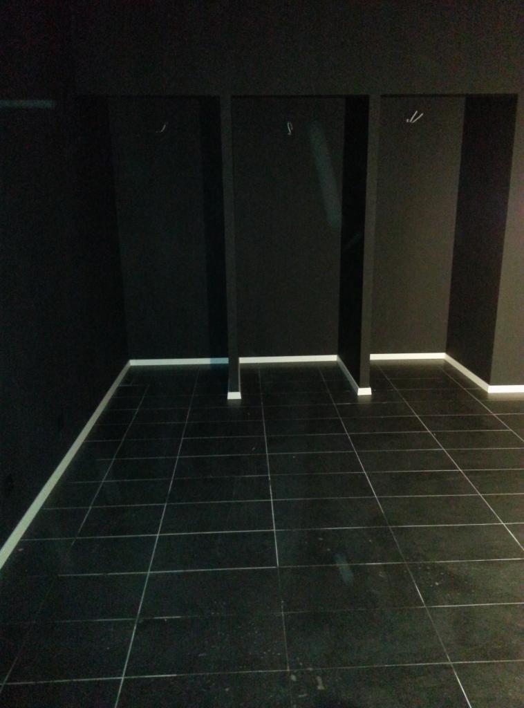 Czarne płytki podłogowe ułożone w szachownicę, czarne ściany z trzema wnękami oddzielonymi filarami i białymi listwami przypodłogowymi w pustym, ciemnym pomieszczeniu.