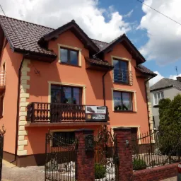 Dwupiętrowy dom z pomarańczową elewacją, brązowymi oknami i dachem, kremowymi detalami architektonicznymi oraz balkonami z metalowymi balustradami, widoczny zza ozdobnego, kutego ogrodzenia...