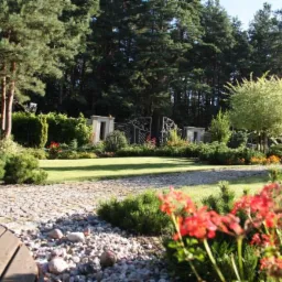 Urokliwy ogród z trawnikiem, kamienną ścieżką, ozdobną bramą i bujną roślinnością, w tym czerwonymi kwiatami na pierwszym planie oraz drzewami w tle.