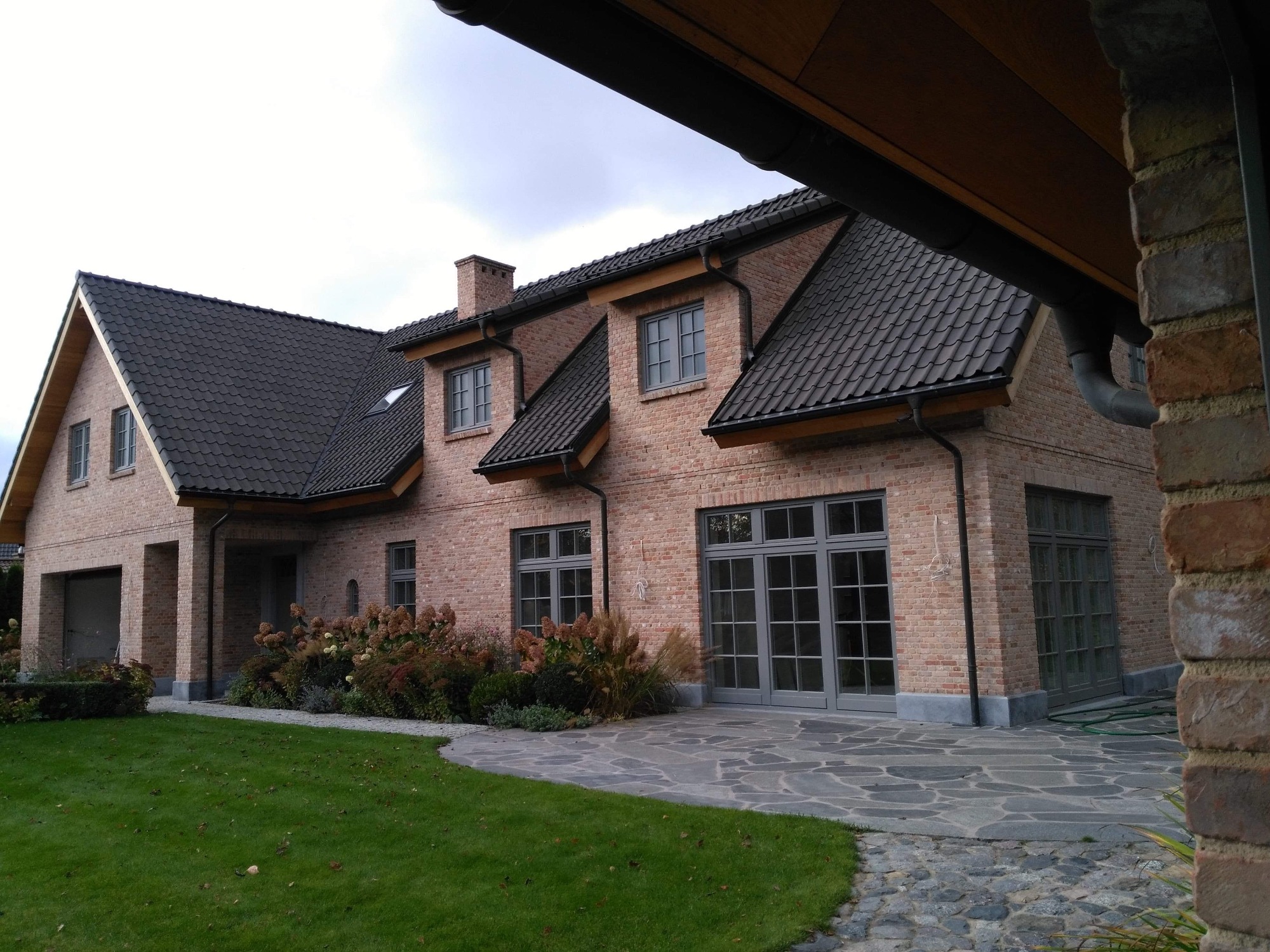 Nowoczesny dom z czerwonej cegły z ciemnoszarą dachówką, szare okna i drzwi balkonowe, kamienny chodnik i trawnik, widok z zewnątrz.