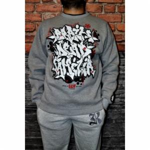 Mężczyzna w szarej bluzie dresowej z dużym, białym, grafitti-podobnym nadrukiem na przodzie, w szarych spodniach dresowych, na tle ściany z cegieł i drewnianych paneli.