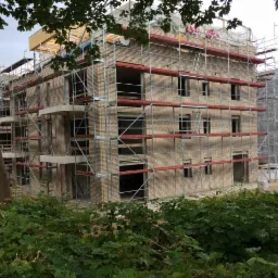 Trzypiętrowy budynek z jasnej cegły w trakcie budowy, otoczony rusztowaniami, widoczny zza zieleni.