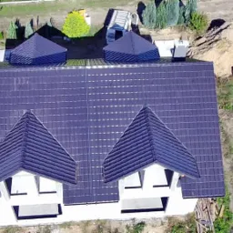 Widok z lotu ptaka na nowy, ciemny dach z blachodachówki na białym domu z czterema małymi wieżyczkami, otoczony zielonym trawnikiem i drzewami.