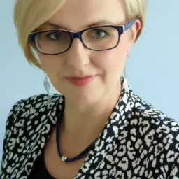 Portret uśmiechniętej kobiety w okularach i wzorzystej marynarce na jasnoniebieskim tle.