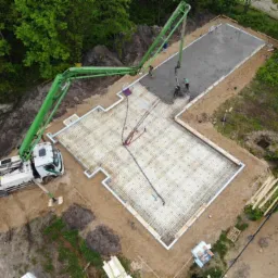 Widok z góry na wylewanie betonu na zbrojenie fundamentów domu jednorodzinnego przy użyciu pompy do betonu z długim ramieniem, w tle zielone drzewa.