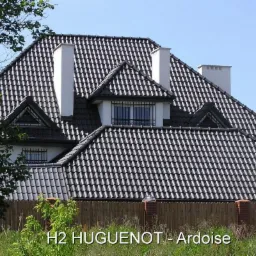 Dom jednorodzinny z ciemnoszarą dachówką karpiówką ułożoną w łuskę, widoczne kominy i okna dachowe, ogrodzenie w tle.