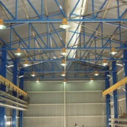 Wnętrze hali produkcyjnej z niebieską konstrukcją stalową, oświetleniem przemysłowym i suwnicą pod stropem.