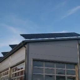 Panele fotowoltaiczne zamontowane na dachu szarego budynku przemysłowego z widocznymi bramami garażowymi i reflektorem, na tle błękitnego nieba.