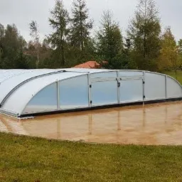 Zadaszenie basenowe z przesuwnymi segmentami na wybrukowanym tarasie, otoczone zielonym trawnikiem i drzewami w tle.