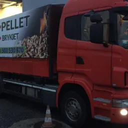 Czerwona ciężarówka z reklamą dostawcy pelletu i brykietu drzewnego z Kędzierzyna-Koźla, zaparkowana przy rampie załadunkowej.