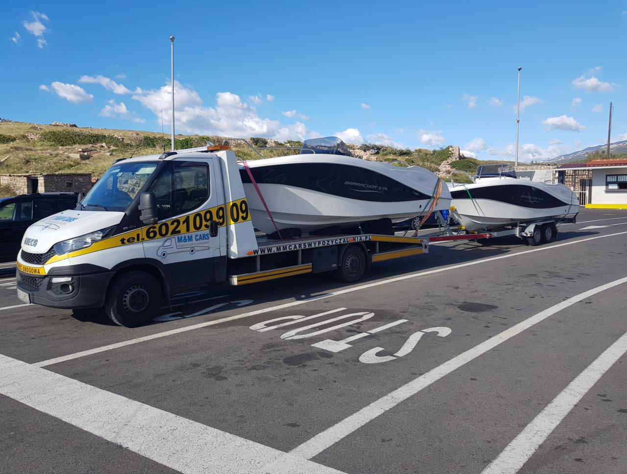 Biała laweta z żółtymi pasami przewozi dwie białe motorówki Barracuda S-45 na przyczepach, na tle błękitnego nieba z chmurami i gór.