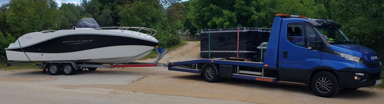 Niebieska laweta Iveco z pomarańczowymi światłami ostrzegawczymi przewozi łódź motorową na przyczepie oraz palety z czarnymi panelami, tło zielone drzewa.