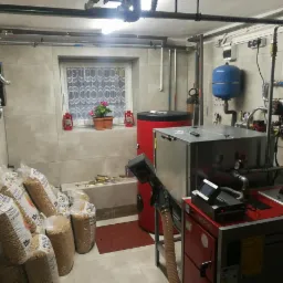 Kotłownia z piecem na pellet Defro, zasobnikiem, workami pelletu, instalacją CO, małym oknem z kwiatami i dekoracyjnymi lampionami.