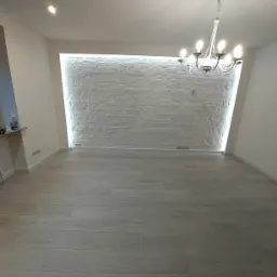 Jasne panele podłogowe w pustym pokoju z białą, podświetloną ścianą z cegły i ozdobnym żyrandolem.