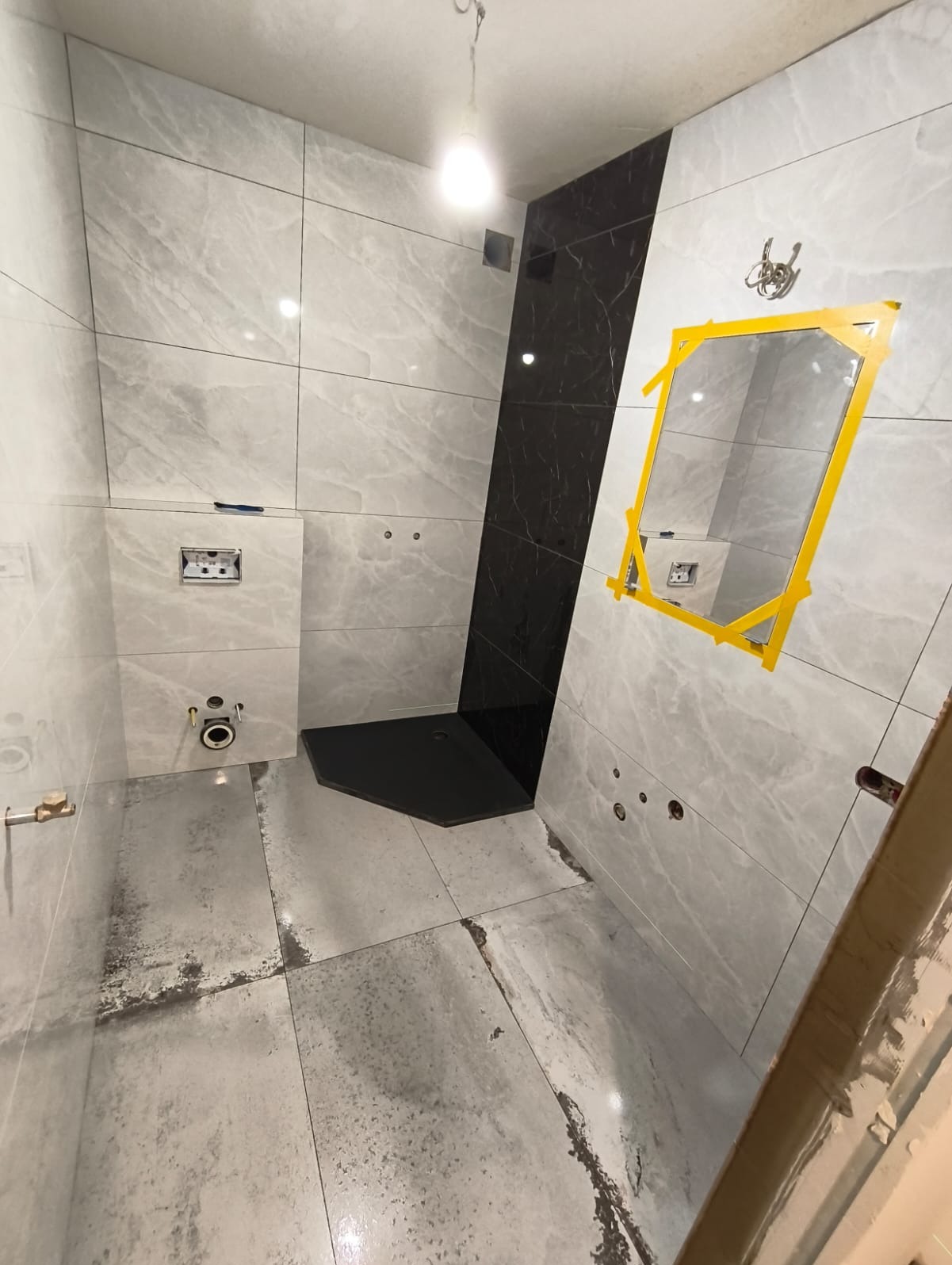Wnętrze łazienki w trakcie remontu: szare płytki imitujące marmur, czarny brodzik, przygotowane miejsce na lustro z żółtą taśmą. Widoczne niedokończone fugi.