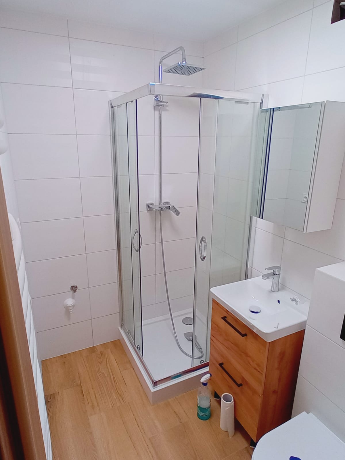 Biała łazienka z kabiną prysznicową typu walk-in, drewnianą szafką pod umywalką i jasną podłogą imitującą drewno. Widok na całe pomieszczenie.