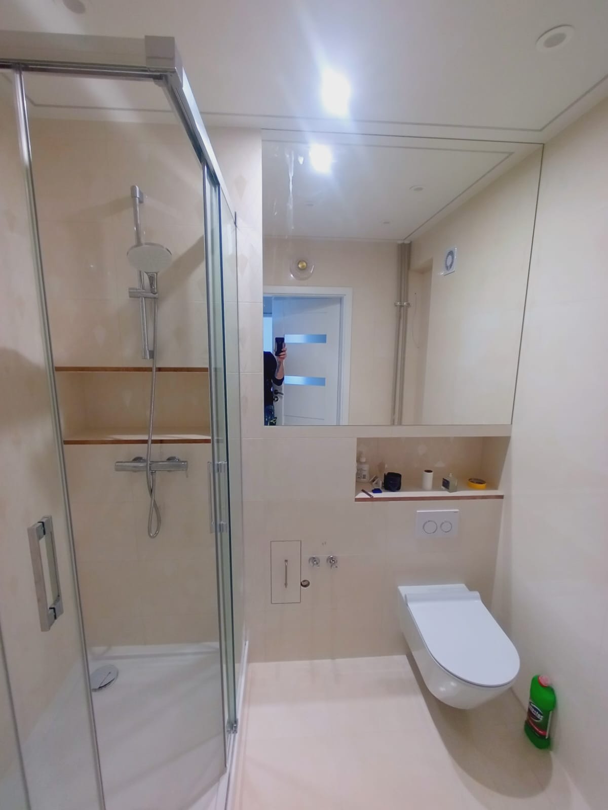 Wykończona łazienka w beżowej tonacji z kabiną prysznicową i podwieszaną toaletą. W lustrze odbija się osoba robiąca zdjęcie.