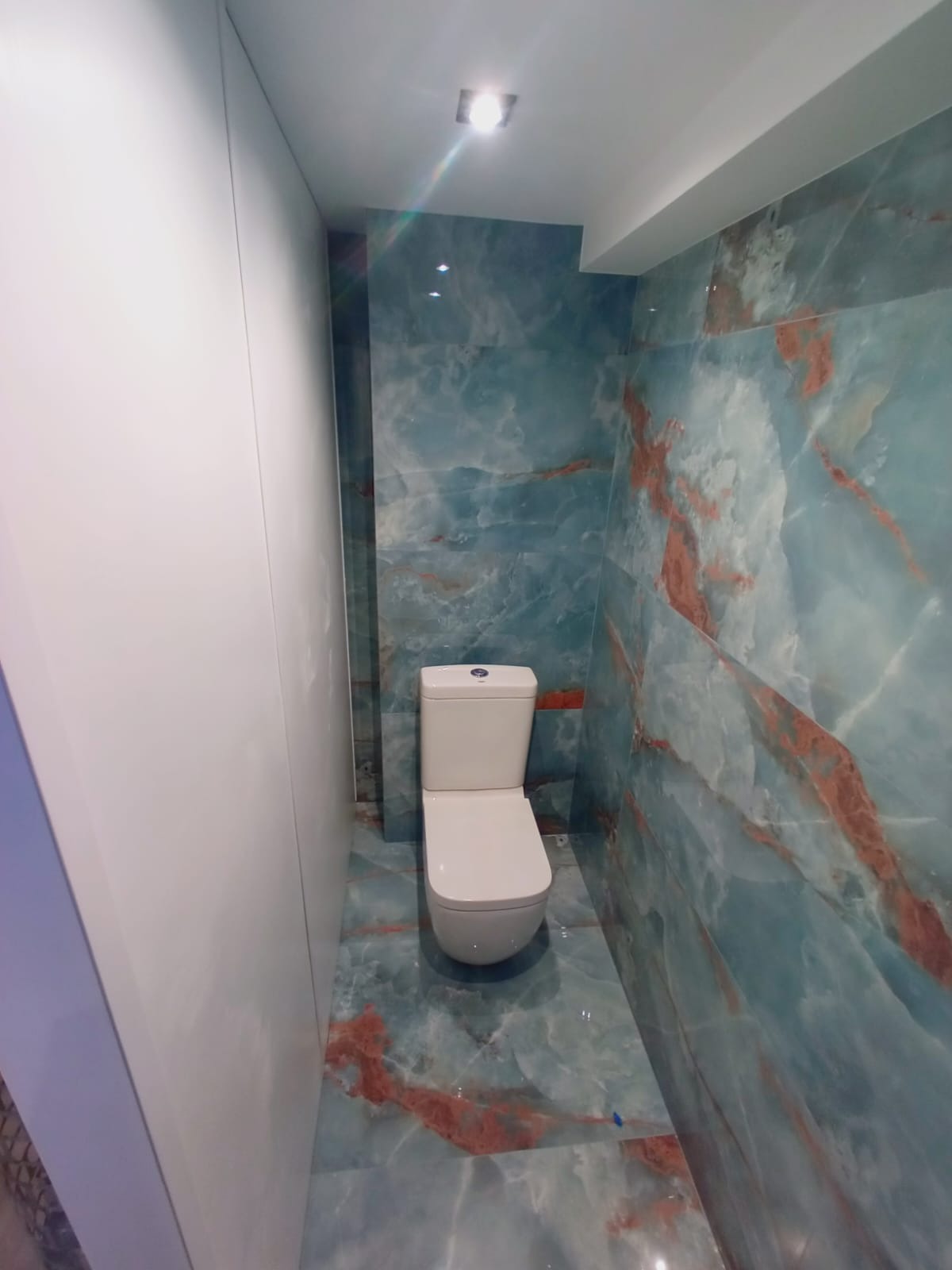 Nowoczesna łazienka z toaletą w kolorze białym i niebiesko-rdzawymi płytkami imitującymi marmur na ścianach i podłodze. Minimalistyczny design, oświetlenie punktowe.