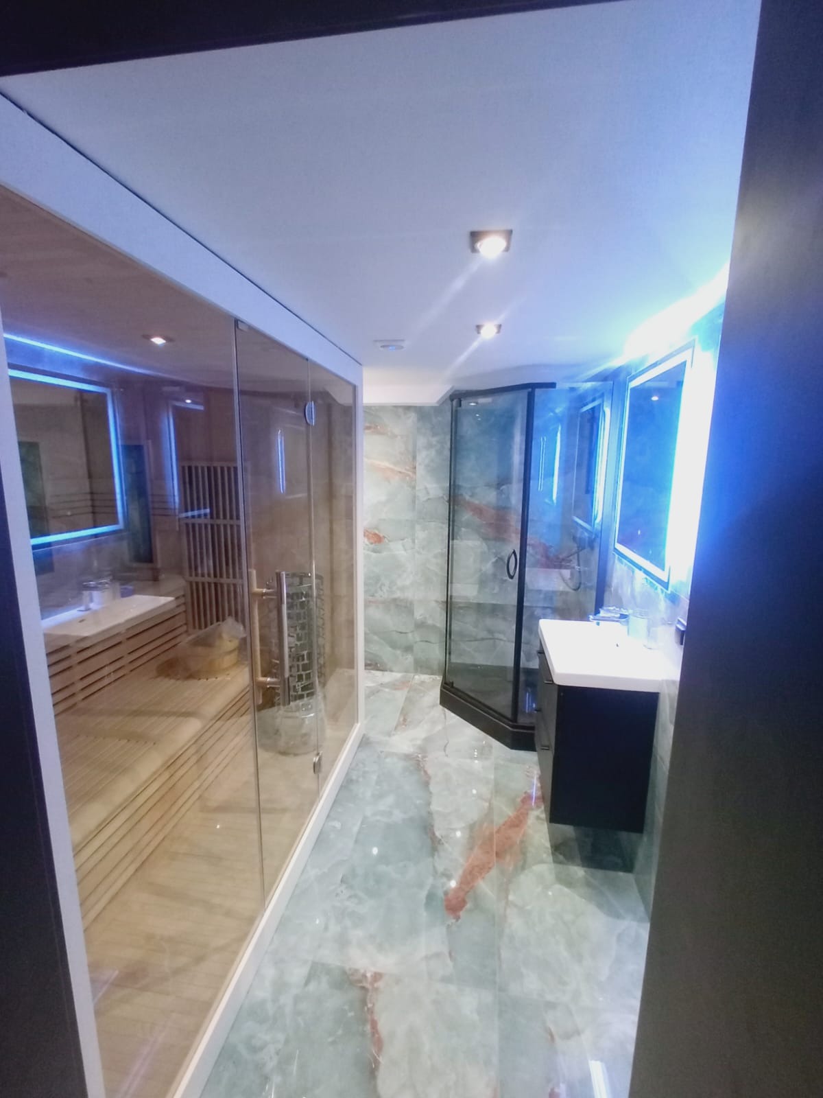 Nowoczesna łazienka z sauną: przeszklona kabina sauny, marmurowe płytki, czarna armatura i lustro z podświetleniem LED. Całość w chłodnej tonacji.