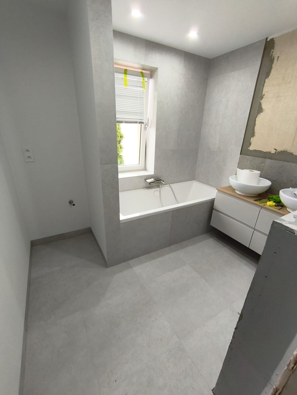 Jasna łazienka w szarościach z wanną, umywalką na szafce i oknem z żaluzjami. Częściowo wykończona ściana. Nowoczesny design, minimalistyczny styl.