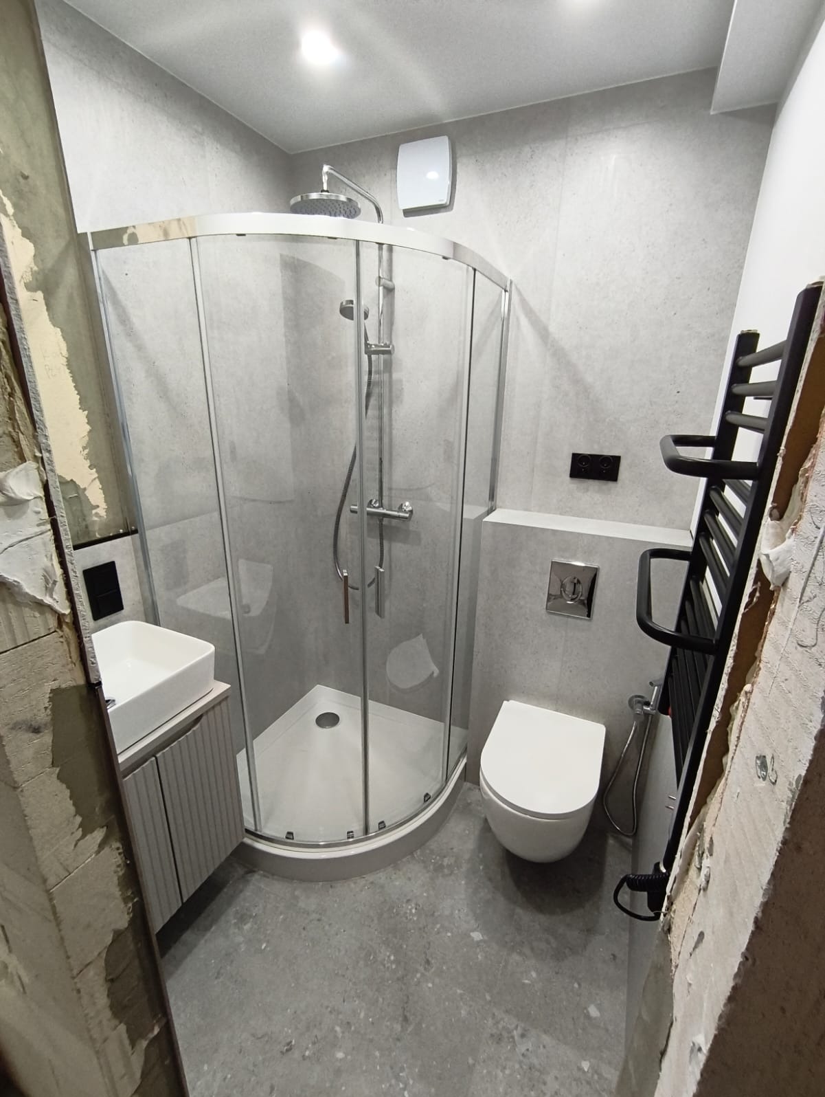 Nowoczesna łazienka w szarościach z kabiną prysznicową narożną, umywalką, toaletą podwieszaną i grzejnikiem drabinkowym. Widoczne ślady prac remontowych.
