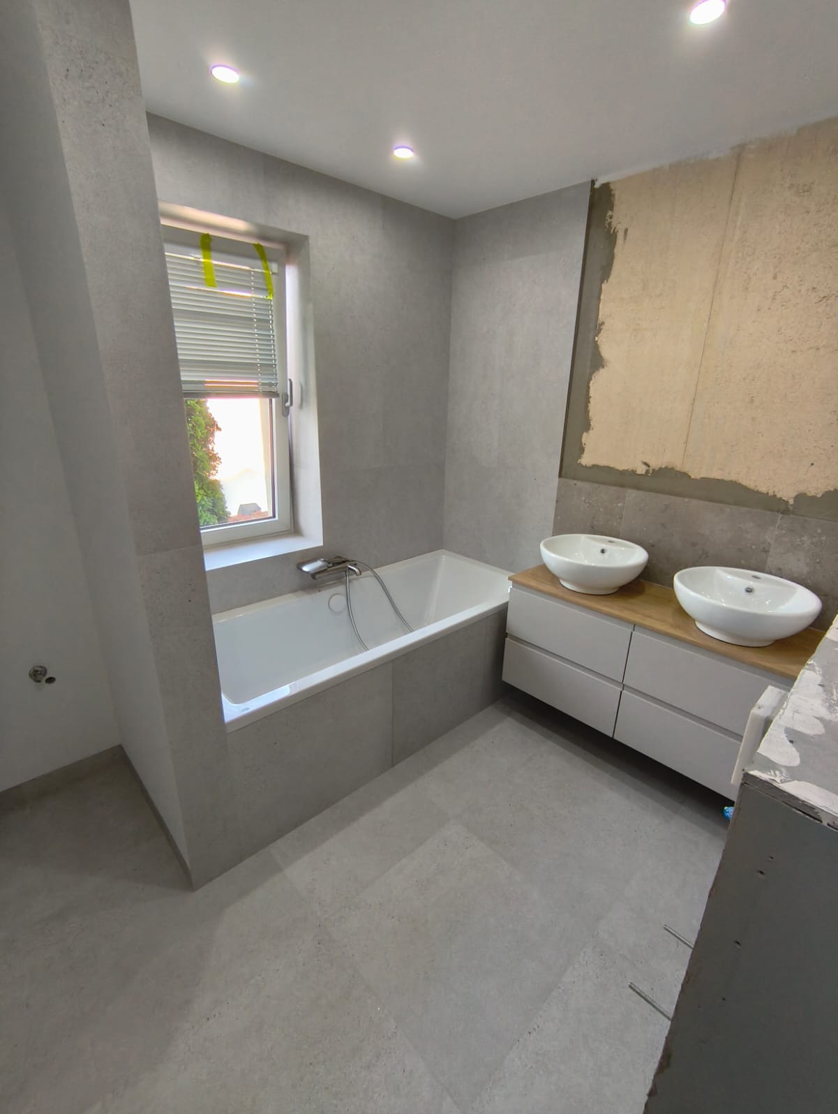 Jasna łazienka w trakcie wykańczania: wanna obudowana płytkami, szafka z umywalkami, szare płytki na ścianach i podłodze. Fragment ściany bez wykończenia.