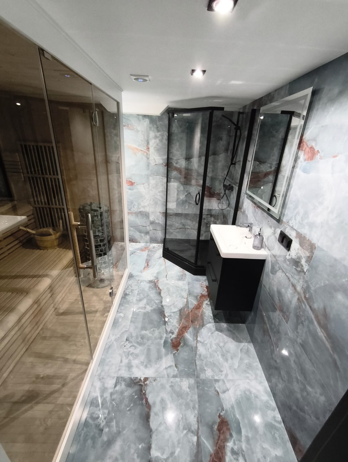 Elegancka łazienka z kabiną prysznicową w czarnej ramie, umywalką na szafce i marmurowymi płytkami. Widoczna sauna z drewnianym wnętrzem za szklaną ścianą.