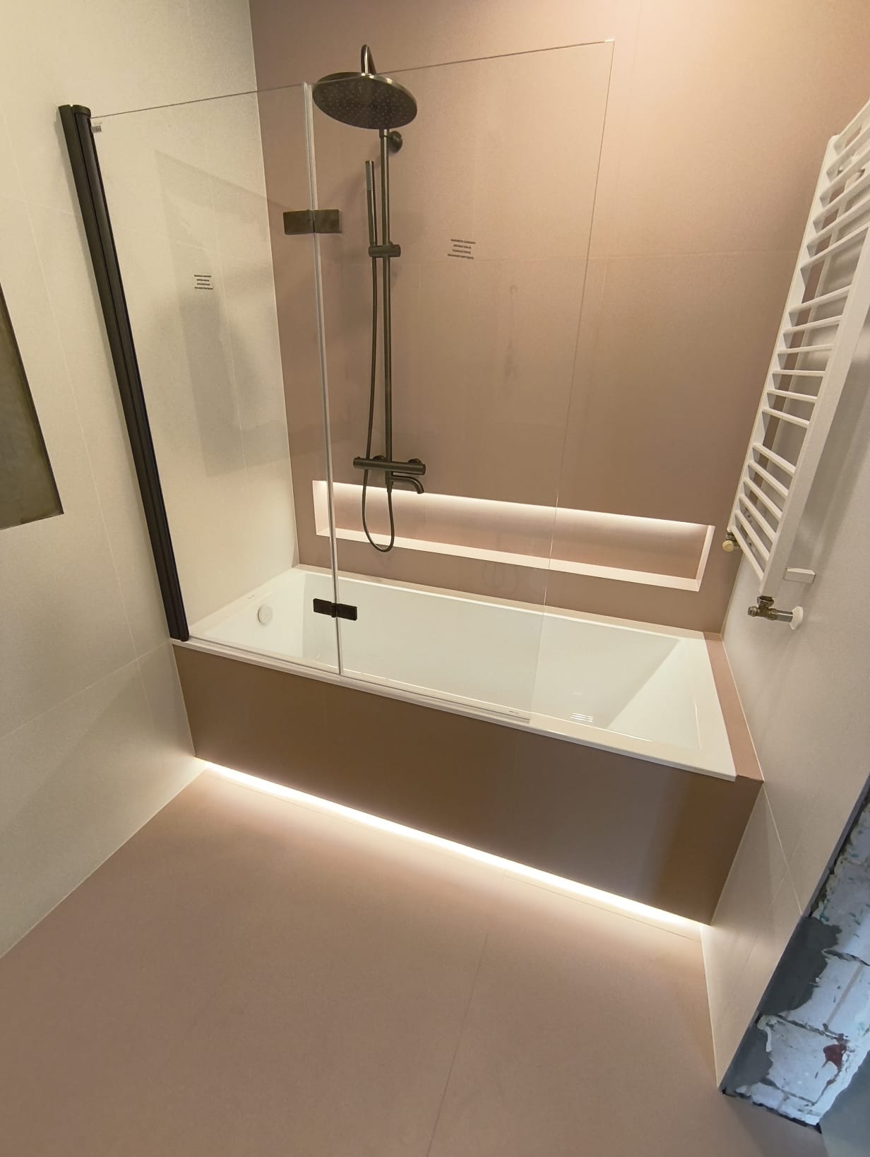 Nowoczesna łazienka z beżowymi płytkami, podświetlaną wanną z parawanem i grzejnikiem drabinkowym. Minimalistyczny design i subtelne oświetlenie tworzą elegancki klimat.