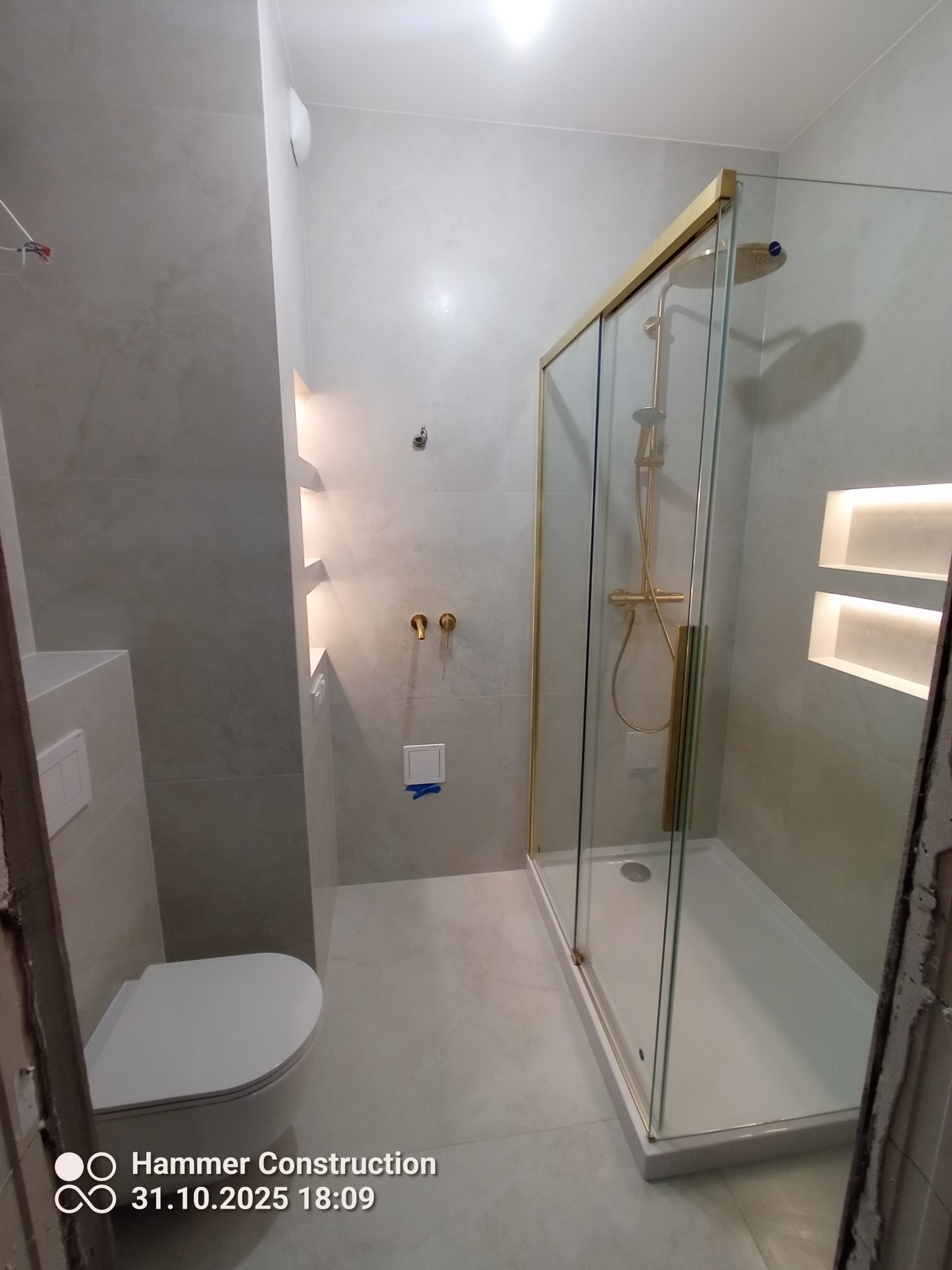 Nowoczesna łazienka z kabiną prysznicową w złotej ramie, beżowe płytki, podświetlane wnęki i wisząca toaleta. Minimalistyczny design z akcentami złota.