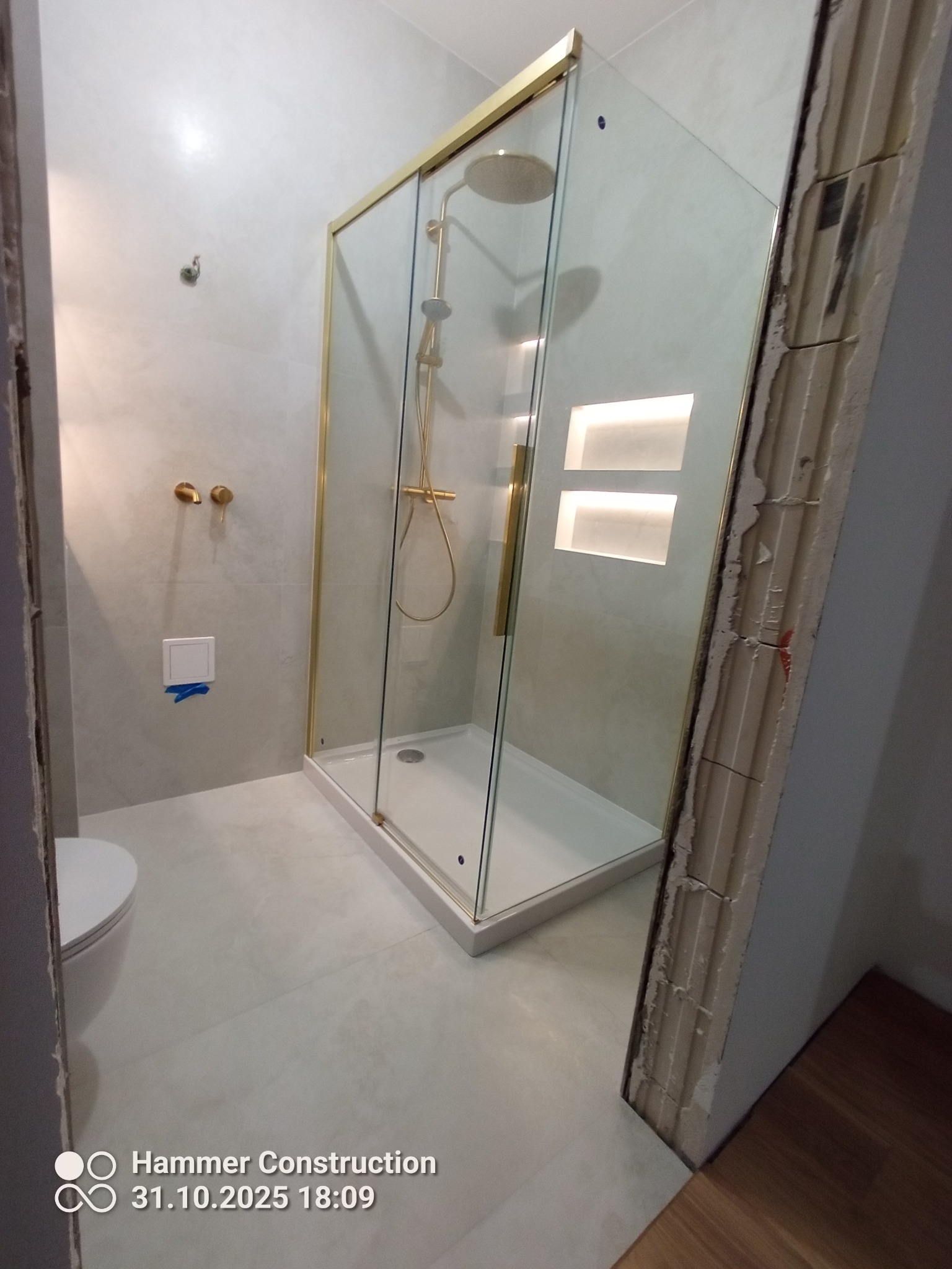 Wykończona łazienka z kabiną prysznicową w złotej ramie, widoczne detale wykończenia ścian i oświetlenie wnękowe. Nowoczesny design.