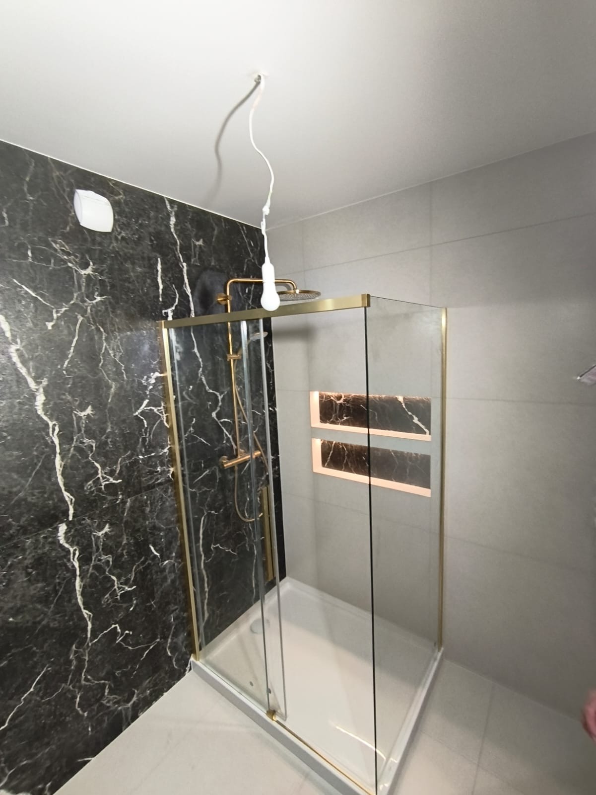 Wykończona łazienka z kabiną prysznicową w złotej ramie, czarną ścianą imitującą marmur i wnękami z podświetleniem. Widoczny biały brodzik i szare płytki.