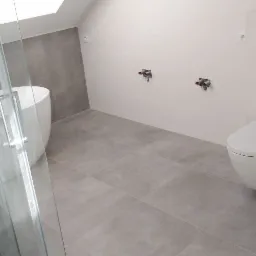 Wykończona łazienka w odcieniach szarości i bieli z wolnostojącą wanną, kabiną prysznicową ze szklanymi drzwiami, podwieszaną toaletą i fragmentem ściany w trakcie wykańczania.