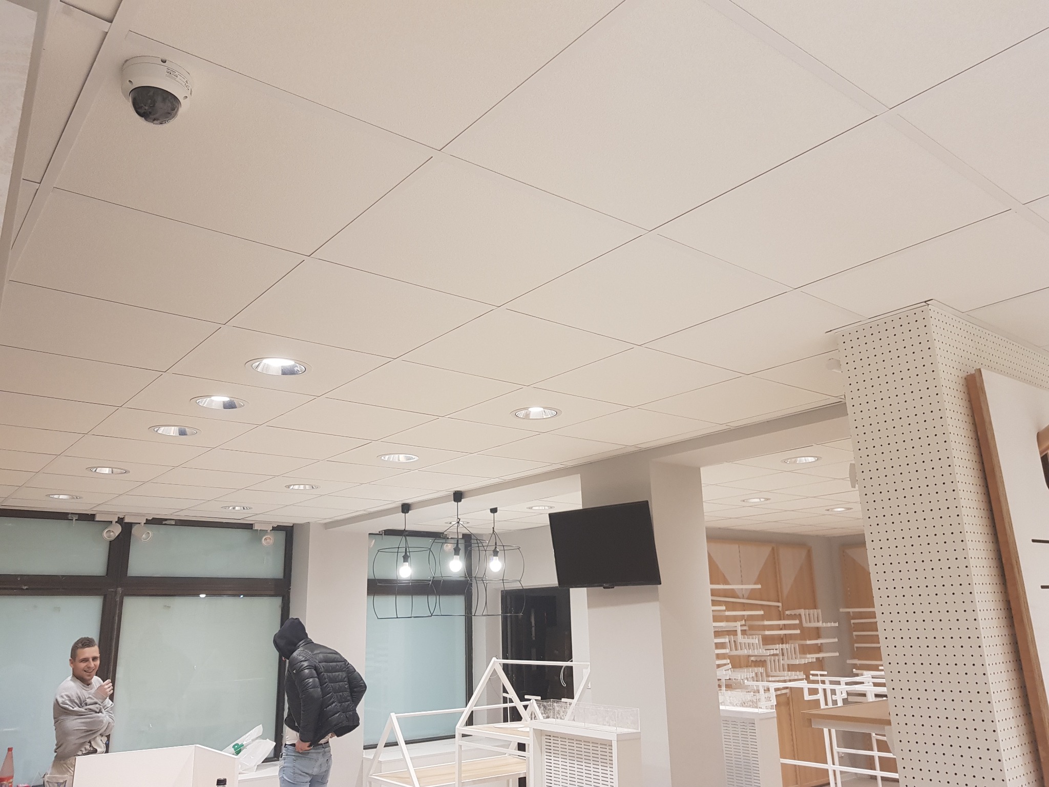 Montaż sufitu podwieszanego kasetonowego z oświetleniem punktowym i kamerą przemysłową w lokalu handlowym, widoczni dwaj mężczyźni podczas prac wykończeniowych.