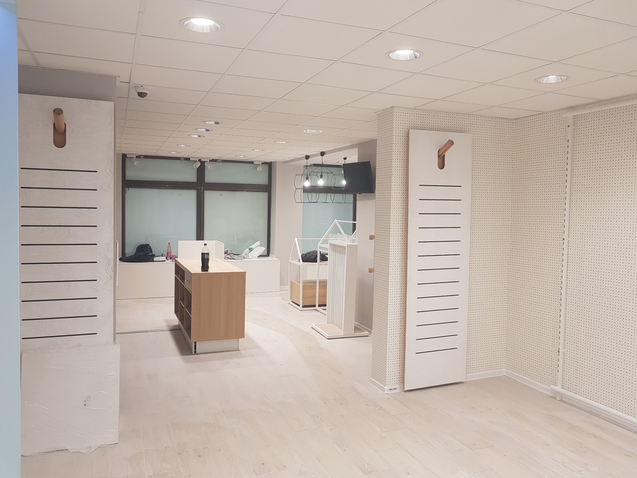 Jasne wnętrze sklepu w trakcie wykańczania, widoczne drewniane podłogi, ściany z panelami perforowanymi, białe elementy wyposażenia i minimalistyczne oświetlenie sufitowe.