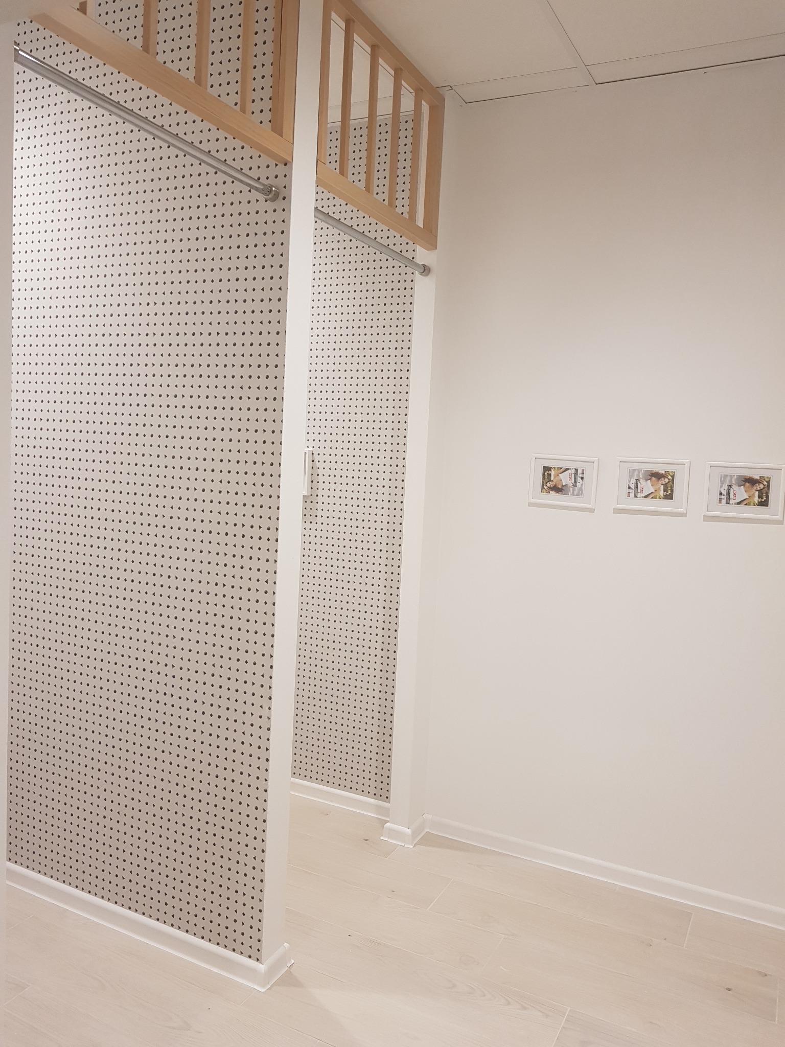 Wykończone wnętrze garderoby z perforowanymi ścianami, drewnianą konstrukcją nośną z drążkiem na ubrania i trzema ramkami ze zdjęciami na ścianie w tle.