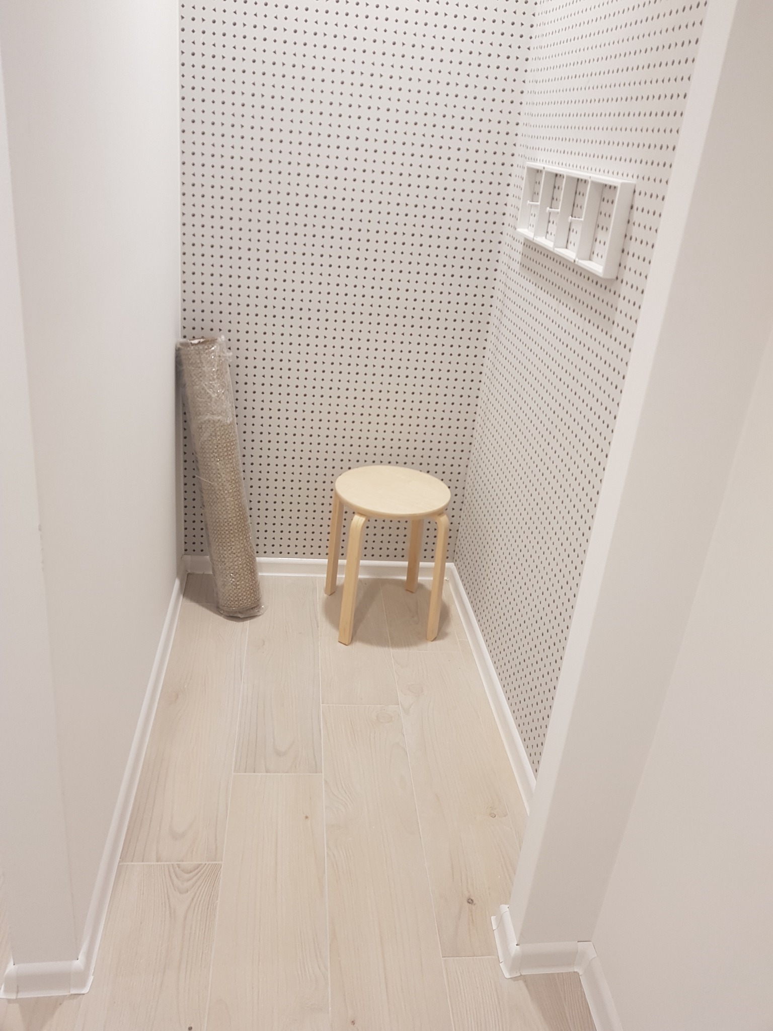 Wąskie pomieszczenie z drewnianą podłogą i ścianami pokrytymi tapetą w drobne kropki, w rogu zwinięty dywan w folii, obok mały drewniany stołek, na ścianie biała półka.