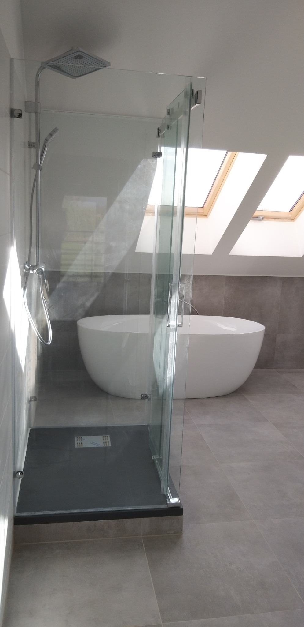 Nowoczesna łazienka na poddaszu z kabiną prysznicową typu walk-in i wolnostojącą wanną owalną, oświetlona naturalnym światłem z okien dachowych.