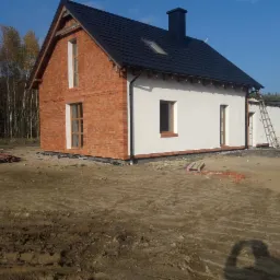 Dwukondygnacyjny dom z częściowo wykończoną elewacją: jedna ściana z cegły, druga otynkowana na biało, czarny dach i komin, drabina oparta o ścianę, teren wokół domu w trakcie prac wykończeniowych.