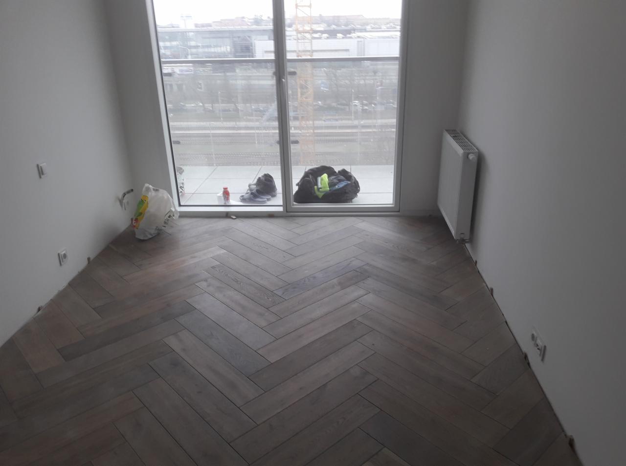 Wnętrze pokoju z ułożoną parkietową podłogą w jodełkę, widoczne niedokończone prace wykończeniowe przy ścianach, okno balkonowe z widokiem na budynek w tle, grzejnik na ścianie, torba i ubrania...