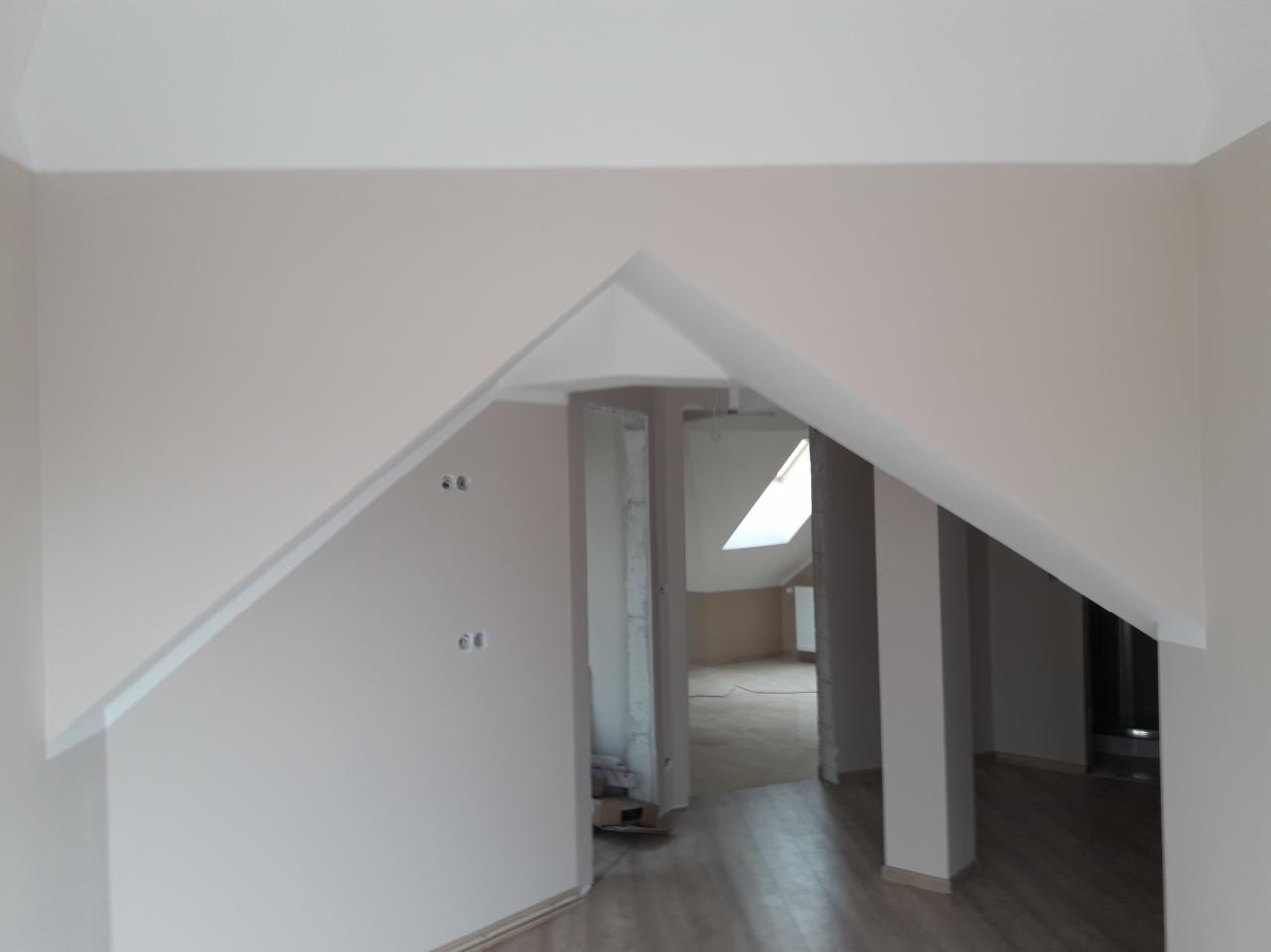 Poddasze po remoncie z nowymi ścianami w odcieniach beżu i bieli, widoczne wystające kable elektryczne i fragment podłogi z paneli, w tle okno dachowe.