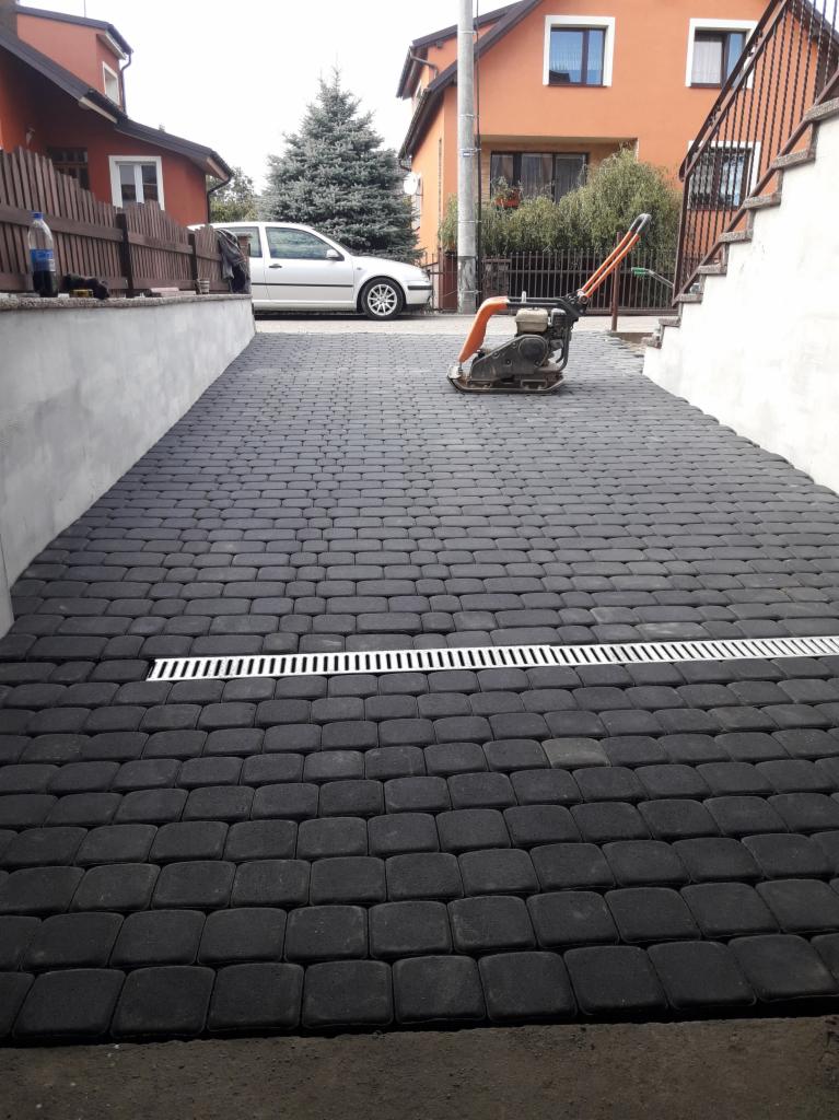 Ułożona, grafitowa kostka brukowa na podjeździe z widocznym odpływem liniowym i zagęszczarką spalinową, domy w tle.