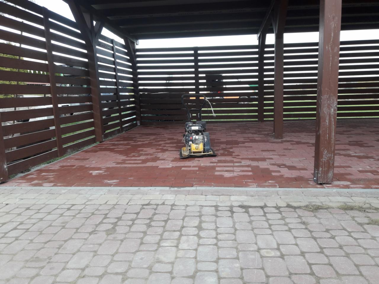 Zagęszczarka spalinowa na mokrej, czerwonej kostce brukowej pod drewnianą konstrukcją z zadaszeniem i ażurowymi ścianami, widok z zewnątrz na ułożoną kostkę.