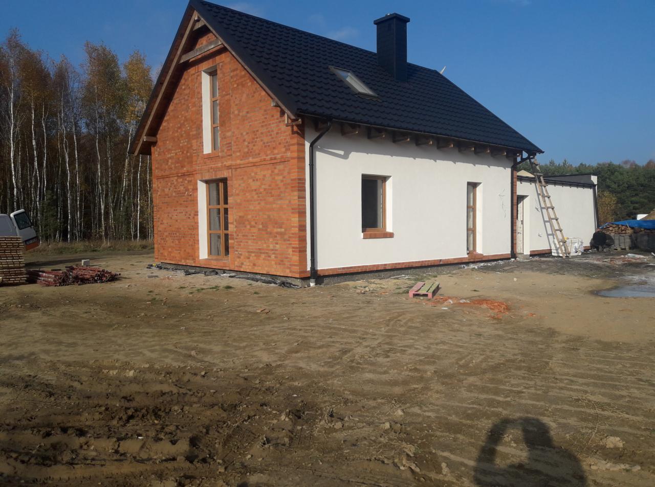 Dwukondygnacyjny dom z częściowo wykończoną elewacją: jedna ściana z cegły, druga otynkowana na biało, czarny dach i komin, drabina oparta o ścianę, teren wokół domu w trakcie prac wykończeniowych.