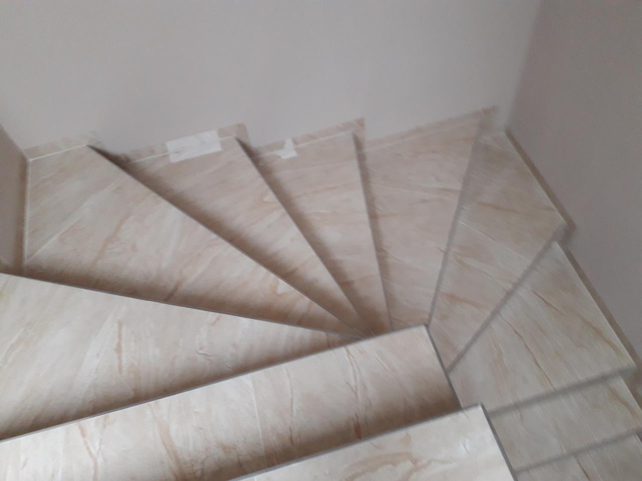 Widok z góry na schody wyłożone płytkami imitującymi marmur w odcieniach beżu i kremu, z widocznymi fragmentami białej ściany i sufitu.